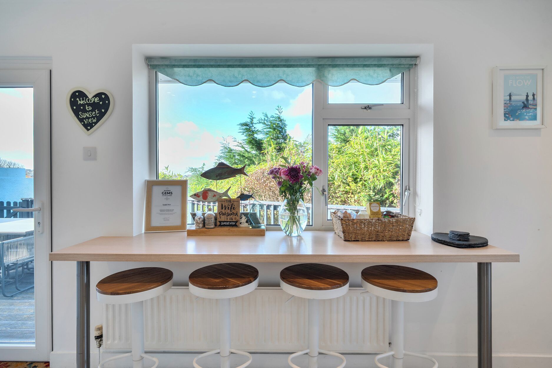 A kitchen counter with a view of the outdoors, a heart-shaped sign, a framed certificate, a vase with flowers, and a basket.