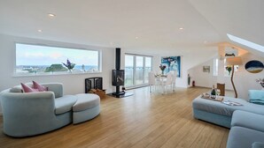 Ein großzügiges Wohnzimmer mit Holzboden, Kamin, Essbereich mit weißen Stühlen und großen Fenstern mit Meerblick.