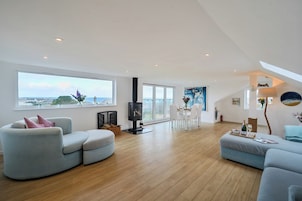 A spacious living room with a wooden floor, a fireplace, a dining area with white chairs, and large windows offering a view of the sea.