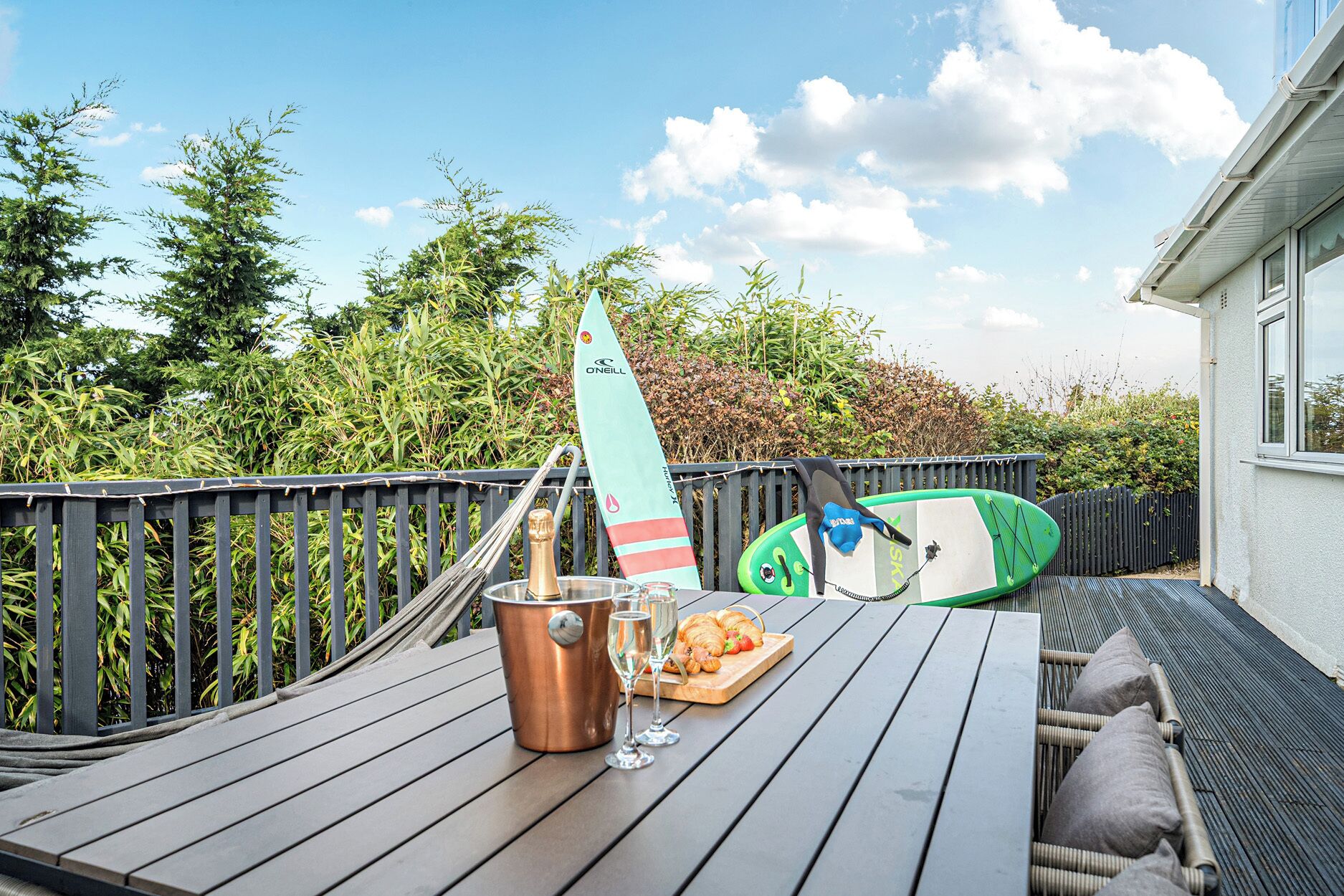 Una terraza de madera con una mesa preparada para una comida, incluyendo una botella de champán, copas y un plato de comida.