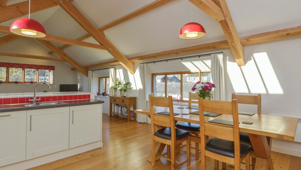 A spacious kitchen with wooden beams, a dining area with a wooden table and chairs, and a large window providing natural light. - Old Armoury Barn (Newton Abbot)
