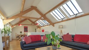 A spacious loft with a high ceiling, exposed wooden beams, a black leather sofa, a wooden coffee table, and skylights. - Old Armoury Barn (Newton Abbot)
