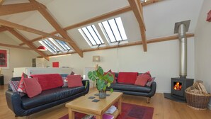 A spacious living room with a black leather sofa, red cushions, a wooden coffee table, and a wood-burning stove. - Old Armoury Barn (Newton Abbot)