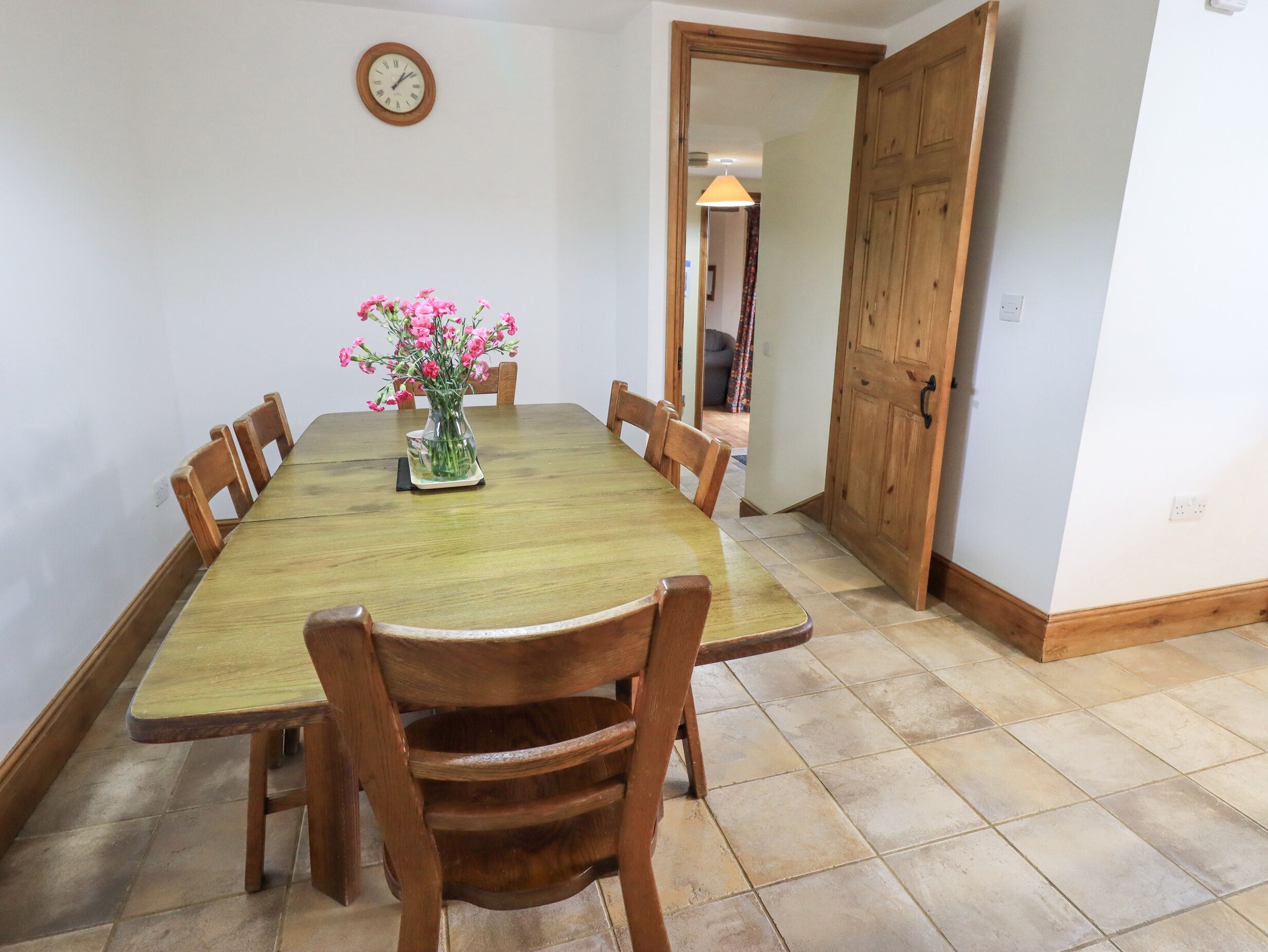 Uma sala de jantar com uma mesa e cadeiras de madeira, um vaso com flores e um relógio na parede.