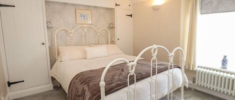 A bedroom with a white metal bed frame, a brown blanket, and a white bedspread. There is a picture on the wall above the bed, and a radiator on the wall to the right.