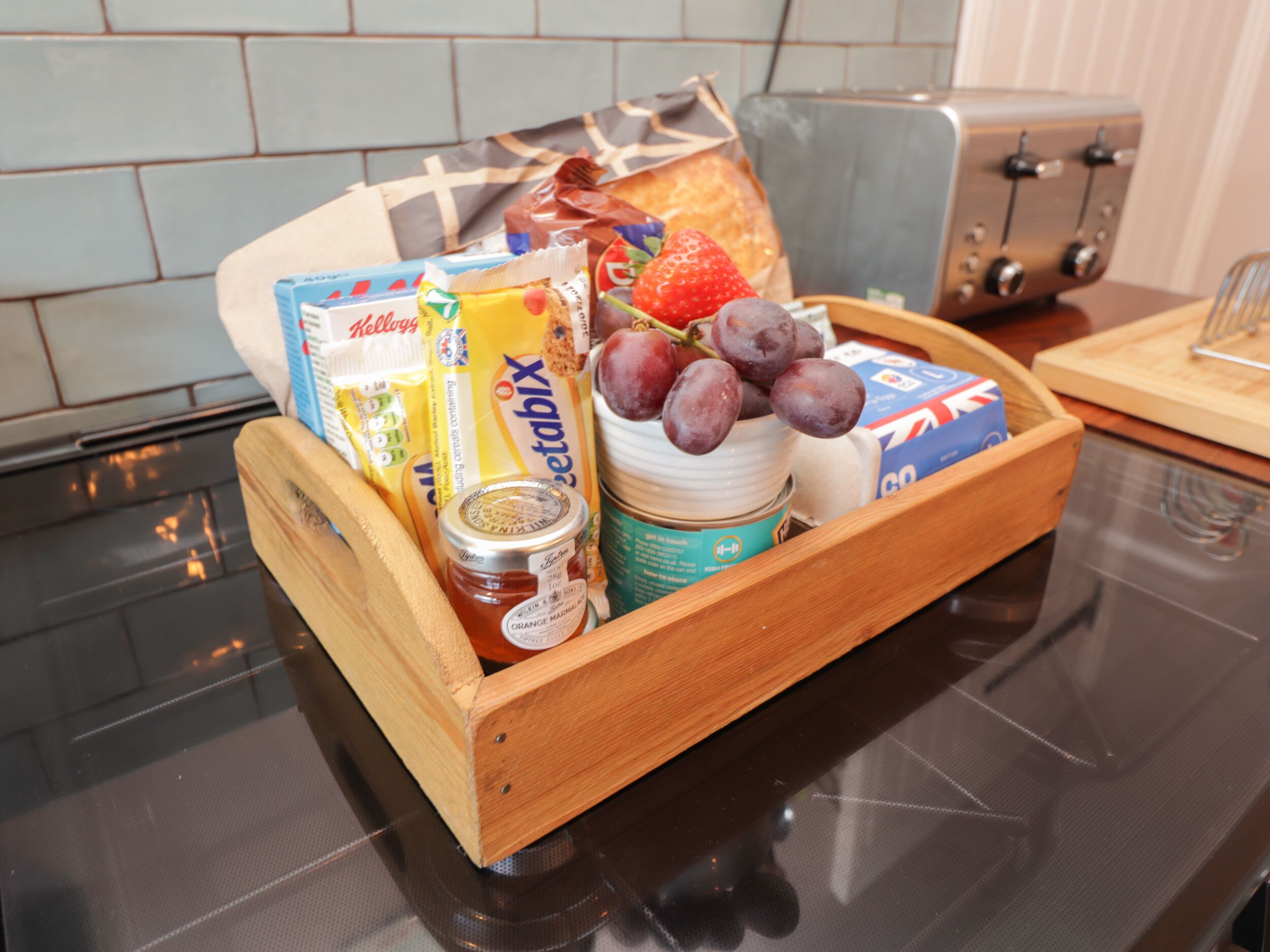 A wooden tray with breakfast items including bread, fruit, cereal, and spreads on a kitchen counter.