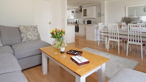 A modern living room with a grey sofa, a wooden coffee table with a vase of flowers, and a dining area with white chairs and a striped tablecloth. - Chyandour (Falmouth)