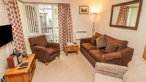 A living room with a brown sofa, a brown armchair, a wooden side table, and a TV on a wooden stand. - Waterhead Apartment B (Ambleside)