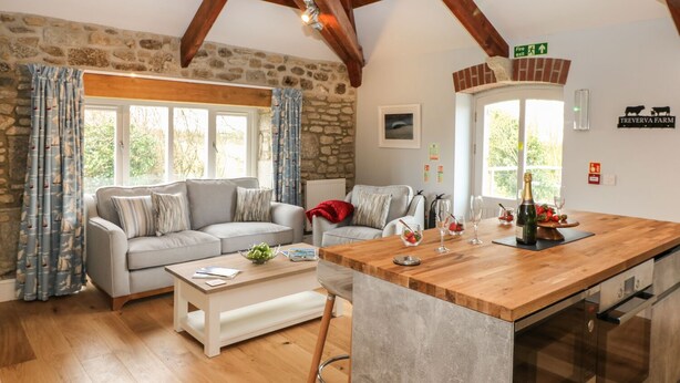 Una sala de estar acogedora con pared de piedra, vigas de madera, un sofá gris, una mesa de centro de madera y una zona de cocina con encimera de madera y electrodomésticos de acero inoxidable.