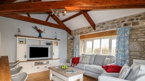 Una sala de estar acogedora con pared de piedra, vigas de madera, televisor, sofá y comedor con sillas.