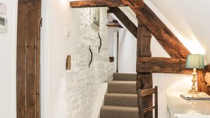 Una escalera con vigas de madera, una pared blanca con detalles de piedra y una mesa con una lámpara.