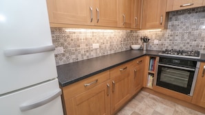 A kitchen with wooden cabinets, a black countertop, a white refrigerator, a stove, and a tiled backsplash. - 12 Kel Avon (Truro)