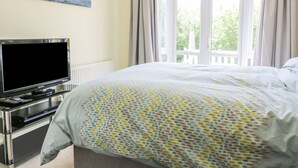 A bedroom with a bed, a television on a stand, a painting on the wall, and a window with curtains. - 12 Kel Avon (Truro)