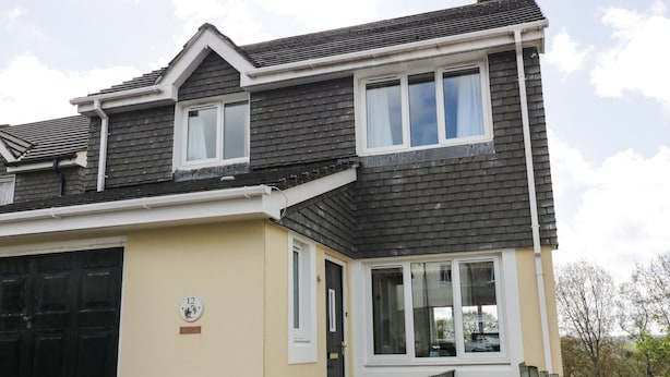 A two-story house with a dark roof, beige walls, and a black garage door. - 12 Kel Avon (Truro)