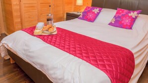 Una cama bien hecha con colcha roja, dos almohadas moradas y una bandeja con cruasanes y café.