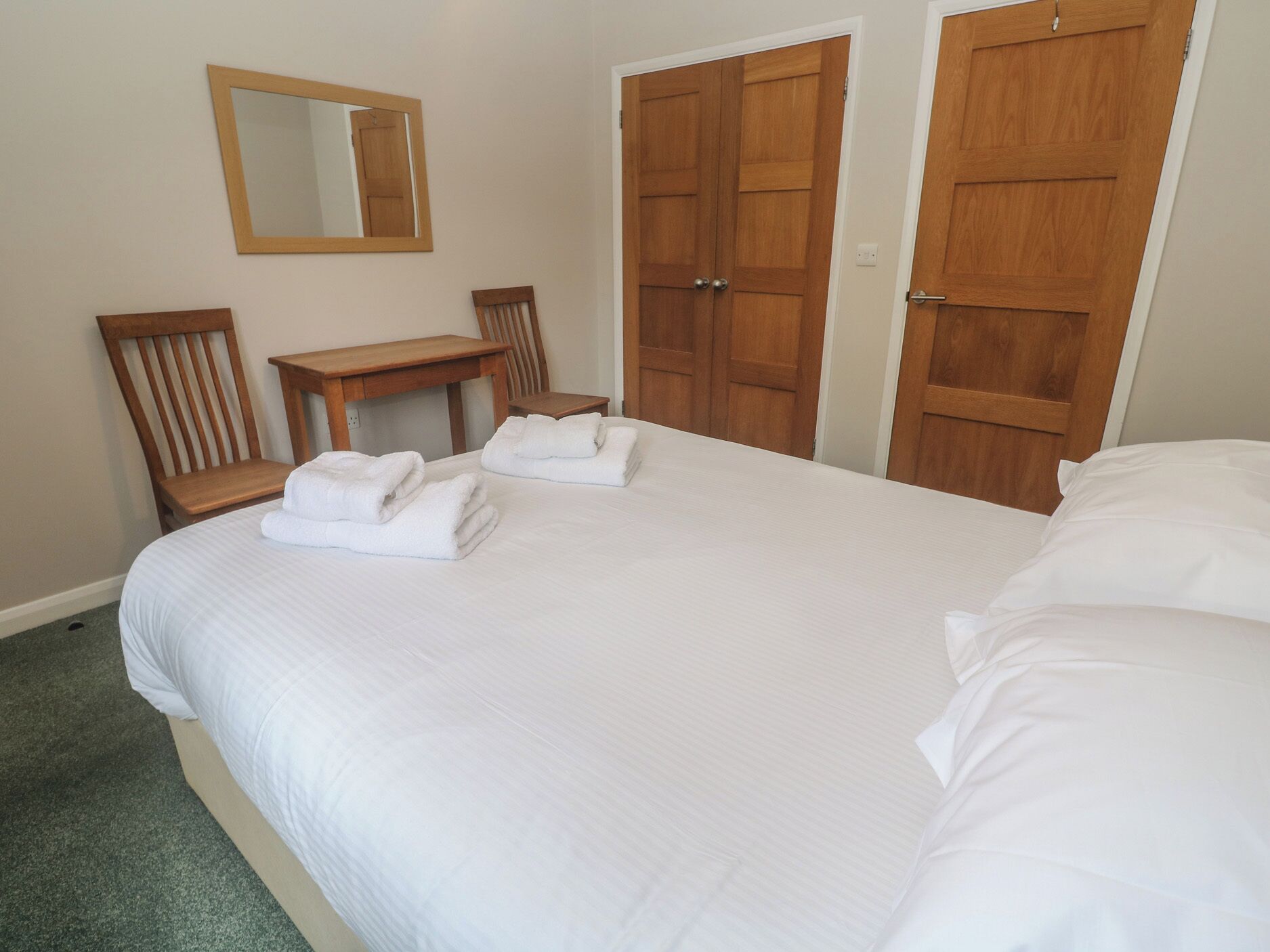 A hotel room with a bed, two wooden doors, a wooden chair, a small table, and a mirror on the wall.