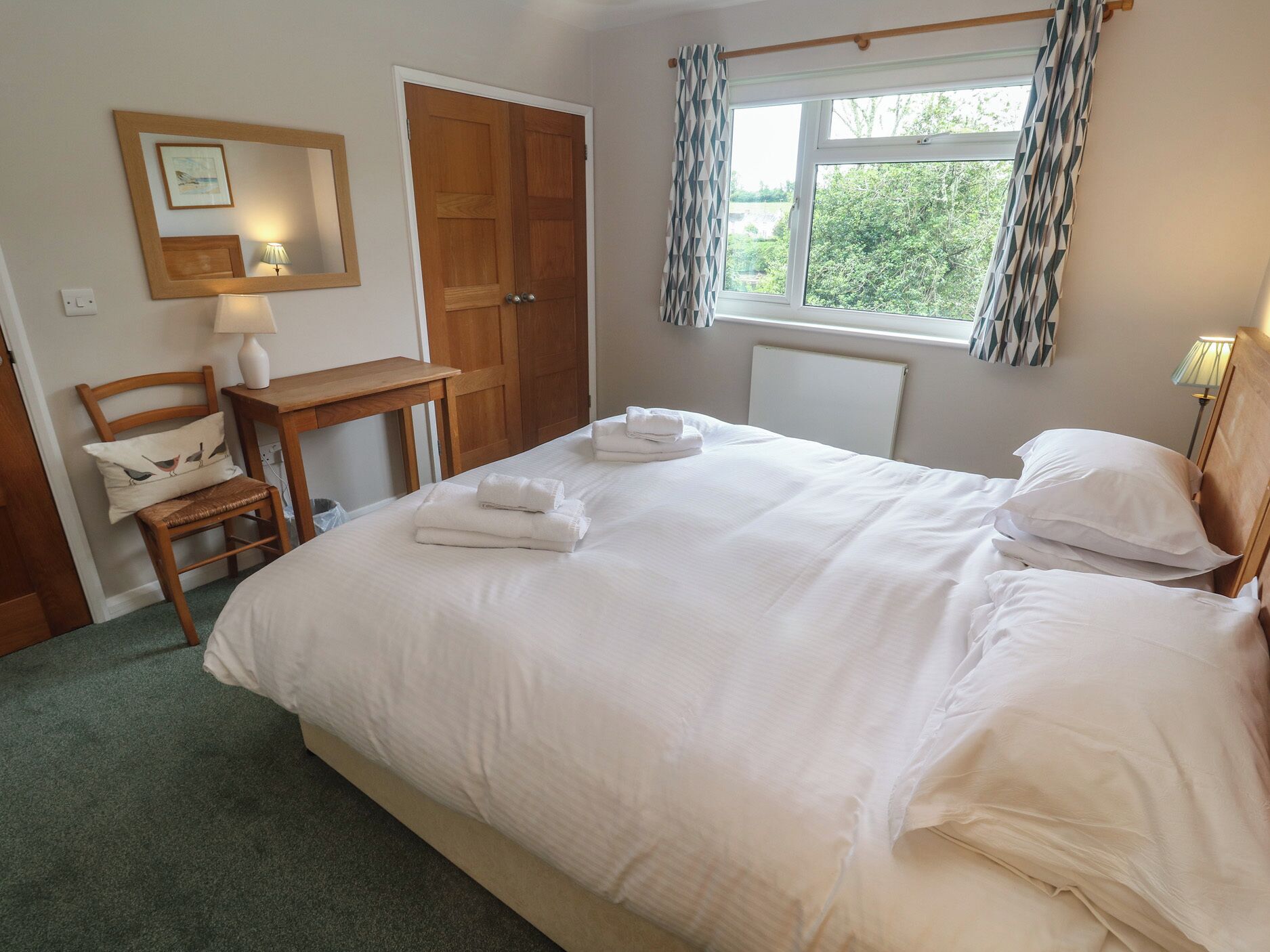 A neatly made bed with white linens and towels, a wooden chair, a desk, a door, and a window with curtains.