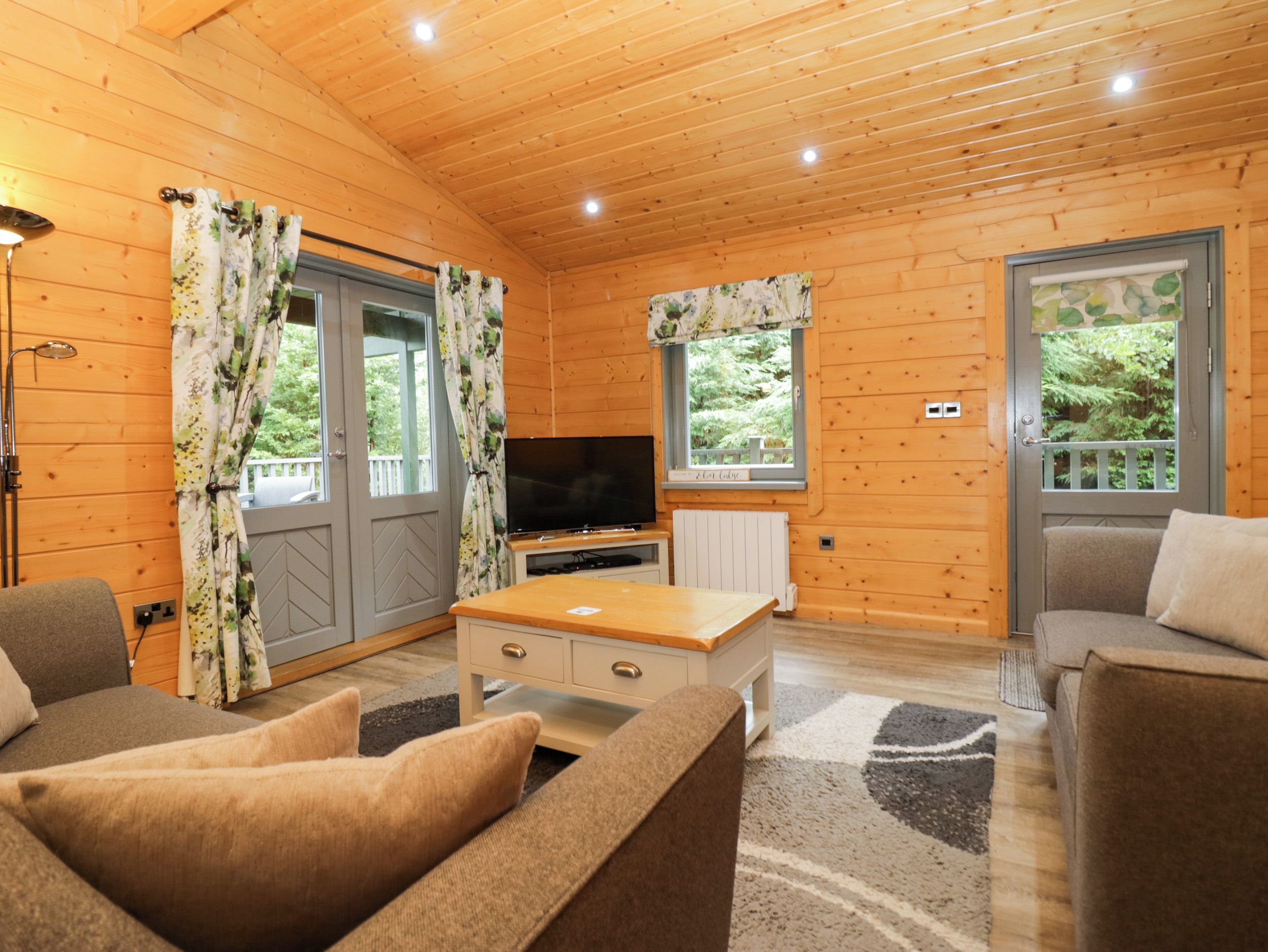 A wooden cabin interior with a sofa, a television, and a window with a view of greenery.