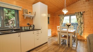 A wooden cabin interior with a kitchen, dining area, and a window with curtains. - Elm Lodge (Ulverston)