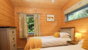 A wooden cabin room with two beds, a dresser, a window with curtains, and a painting on the wall. - Elm Lodge (Ulverston)