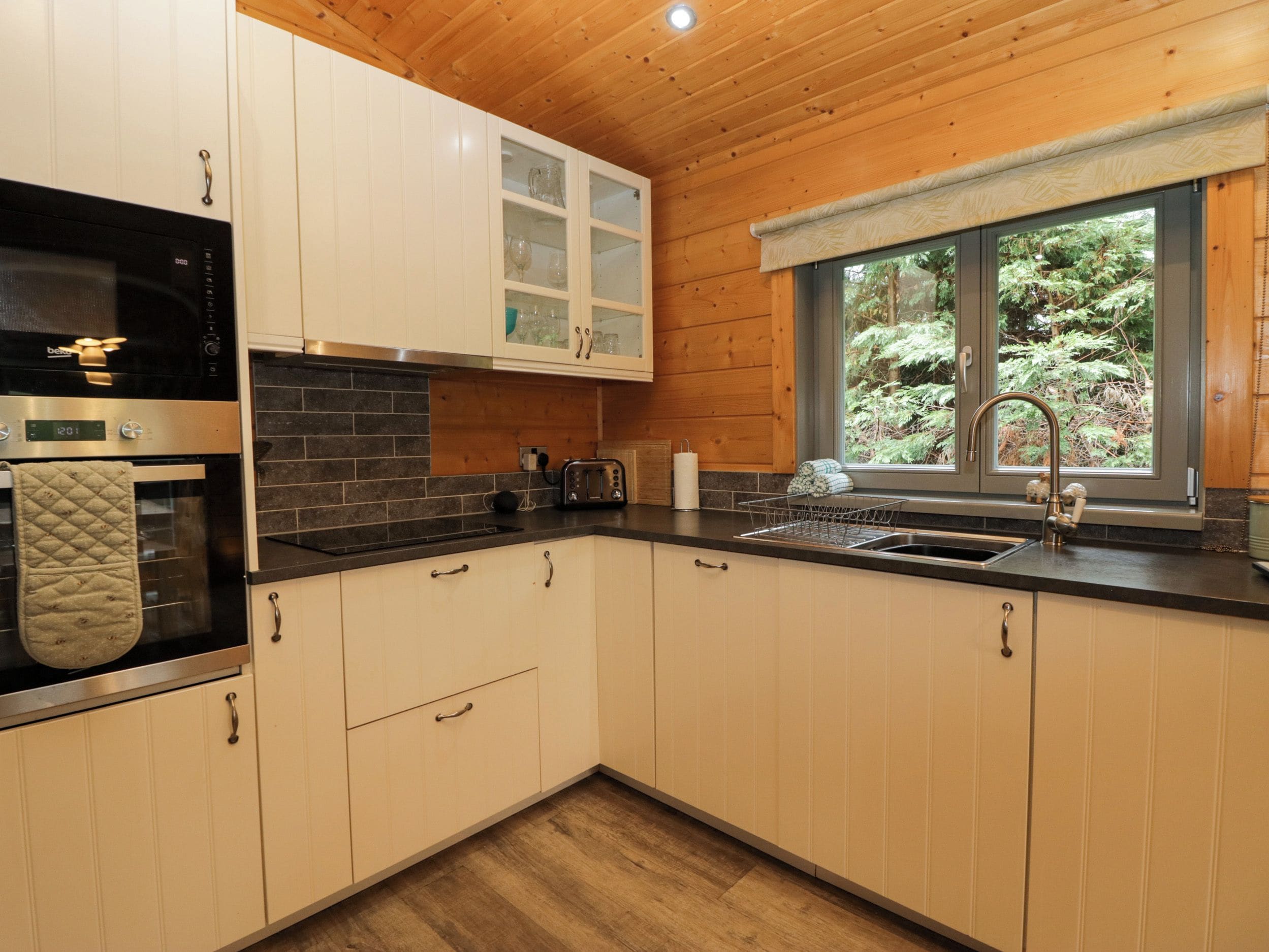 A modern kitchen with wooden cabinets, a black countertop, a stainless steel oven, and a window with a view of greenery.