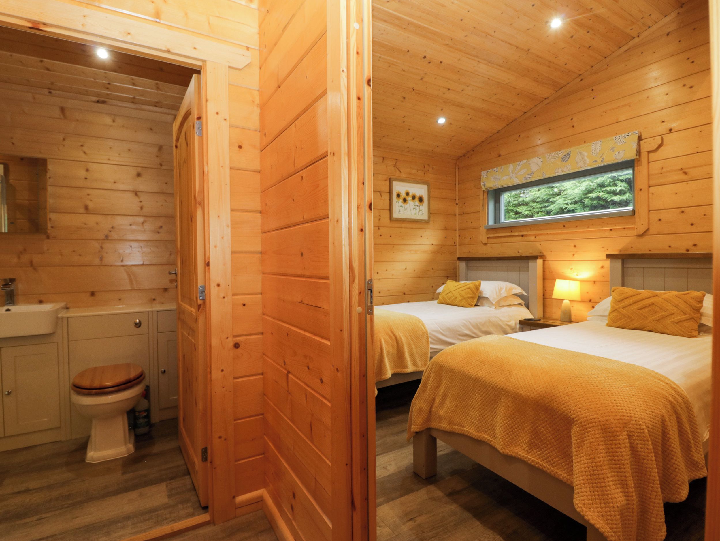 Une cabane en bois avec une salle de bain, deux lits avec du linge de lit jaune et une fenêtre donnant sur un paysage verdoyant.