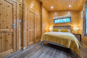 A wooden cabin room with a bed, two bedside lamps, and a window above the bed. - Elm Lodge (Ulverston)