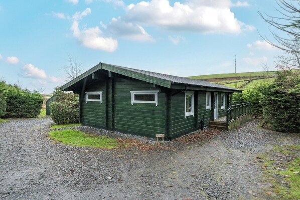A green wooden building with a sloped roof, a porch, and a gravel driveway. - Elm Lodge (Ulverston)