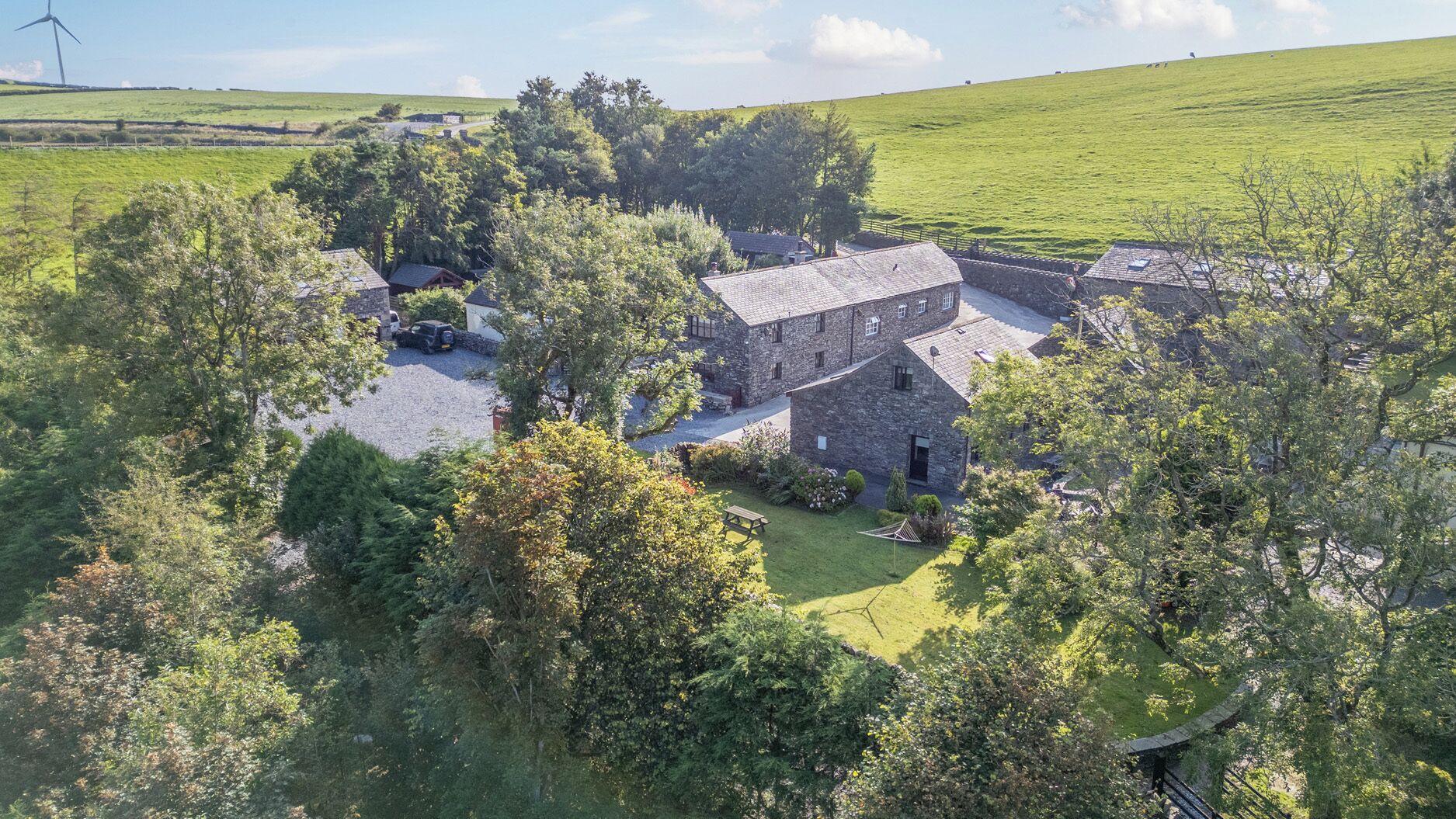 A stone farmhouse with a gravel driveway, surrounded by lush greenery and a wind turbine in the background.