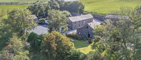 A stone farmhouse with a gravel driveway, surrounded by lush greenery and a wind turbine in the background.