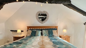 A bedroom with a wooden bed, two bedside tables with lamps, a heart-shaped mirror on the wall, and string lights above the bed.
