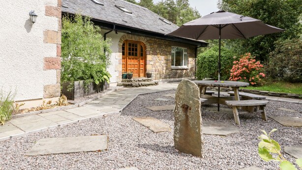 Une maison en pierre avec une porte en bois, une terrasse avec une table et un parasol, et un chemin de gravier.