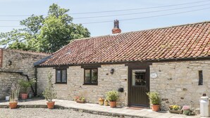 Une petite maison en pierre avec un toit de tuiles, une porte en bois et des plantes en pot devant l’entrée.