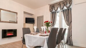 Comedor con mesa dispuesta para cuatro personas, un televisor de pantalla plana sobre la chimenea y una ventana con cortinas.