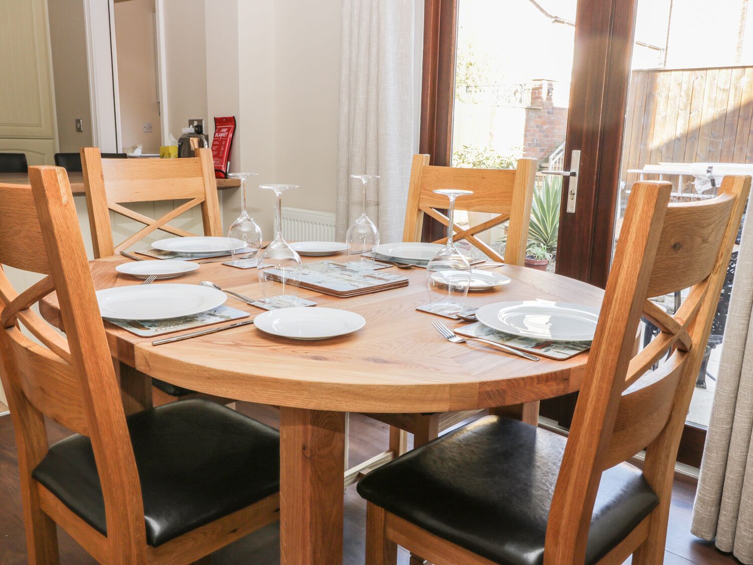 A round wooden dining table set for four with black cushioned chairs, surrounded by a wooden fence and a brick wall.