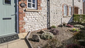 A stone building with a green door, a small window, and a stone pathway leading to a garden with various plants. - Pebble Cottage Dunnscroft (Bridlington)
