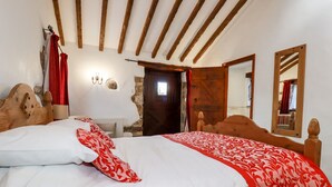 Un dormitorio con techo de madera, una cama grande con ropa blanca y roja, pared de piedra y una puerta de madera.