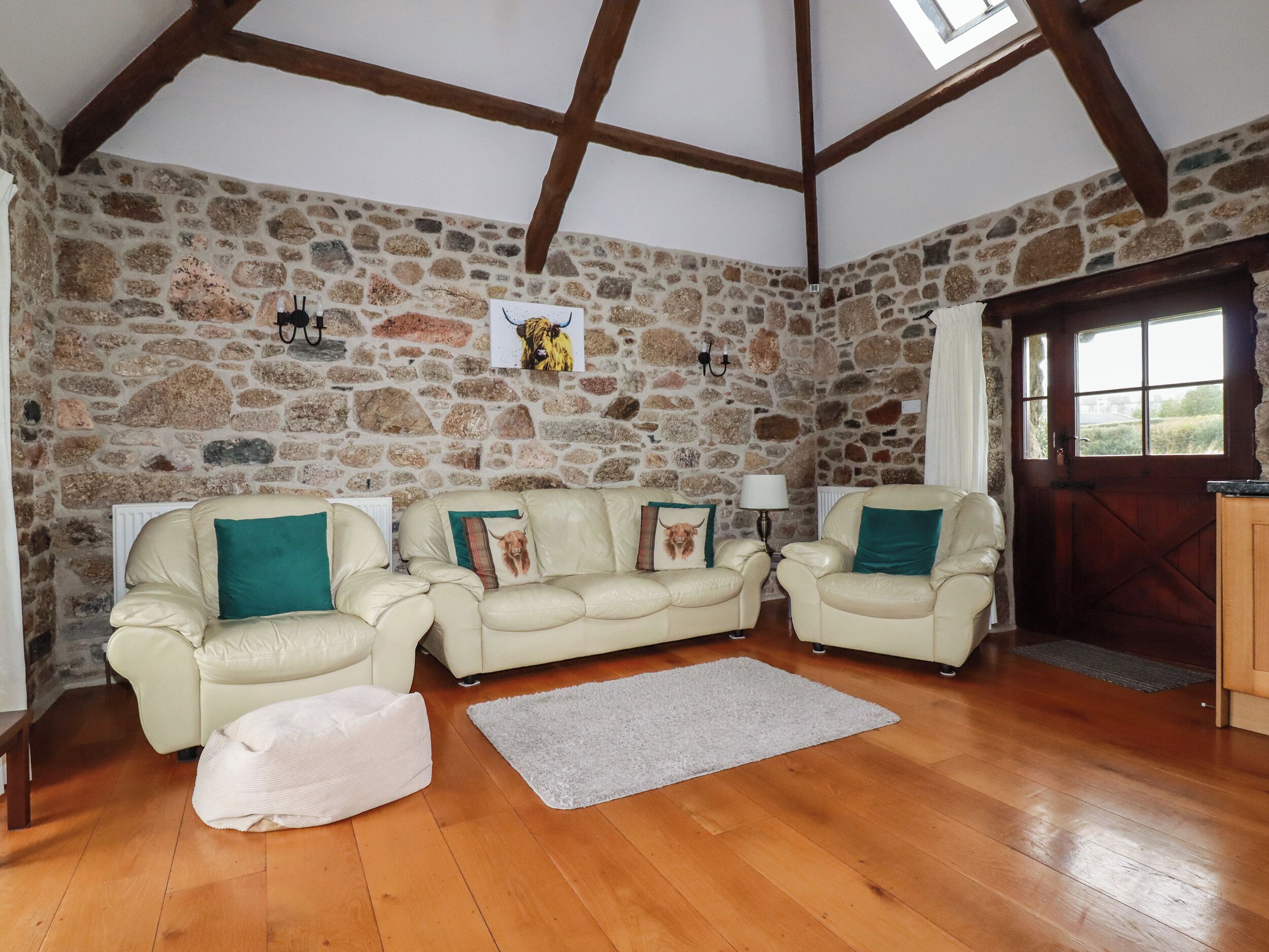 A cozy living room with stone walls, wooden beams, and a stone fireplace. There are two cream-colored sofas with green cushions and a white pouf.