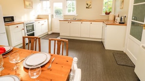 A kitchen with white cabinets, a wooden dining table set for two, and a view of the garden through the window. - Coverdale Cottage (Leyburn)