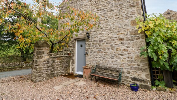 A stone building with a door, a bench, and a tree in front. - Coverdale Cottage (Leyburn)