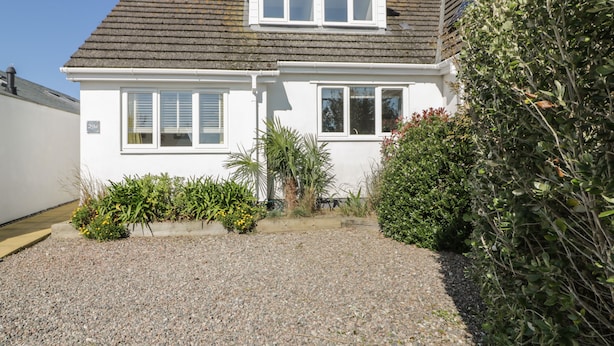 A white two-story house with a gravel driveway and a garden with various plants and shrubs. - Offshore (Newquay)