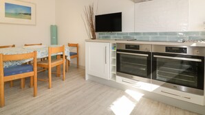 A modern kitchen with two ovens, a built-in TV, and a dining area with wooden chairs and a table. - Offshore (Newquay)