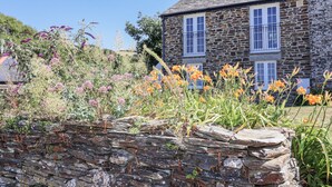 Une maison en pierre avec un toit de tuiles, entourée d’un jardin où poussent différentes sortes de fleurs et bordée d’un mur en pierre.