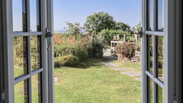 On peut apercevoir, à travers des portes vitrées ouvertes, un jardin agrémenté d’un mur de pierre, d’une pergola en bois et d’une grande variété de plantes et de fleurs.
