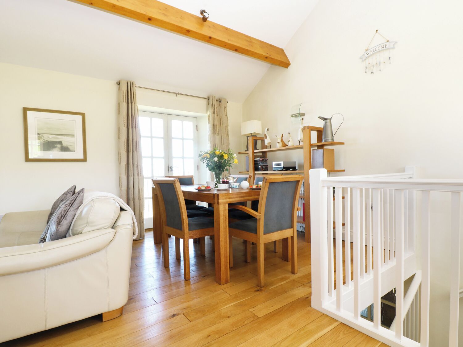 A cozy living room with a white sofa, wooden dining table, and chairs. A white crib is visible in the foreground.