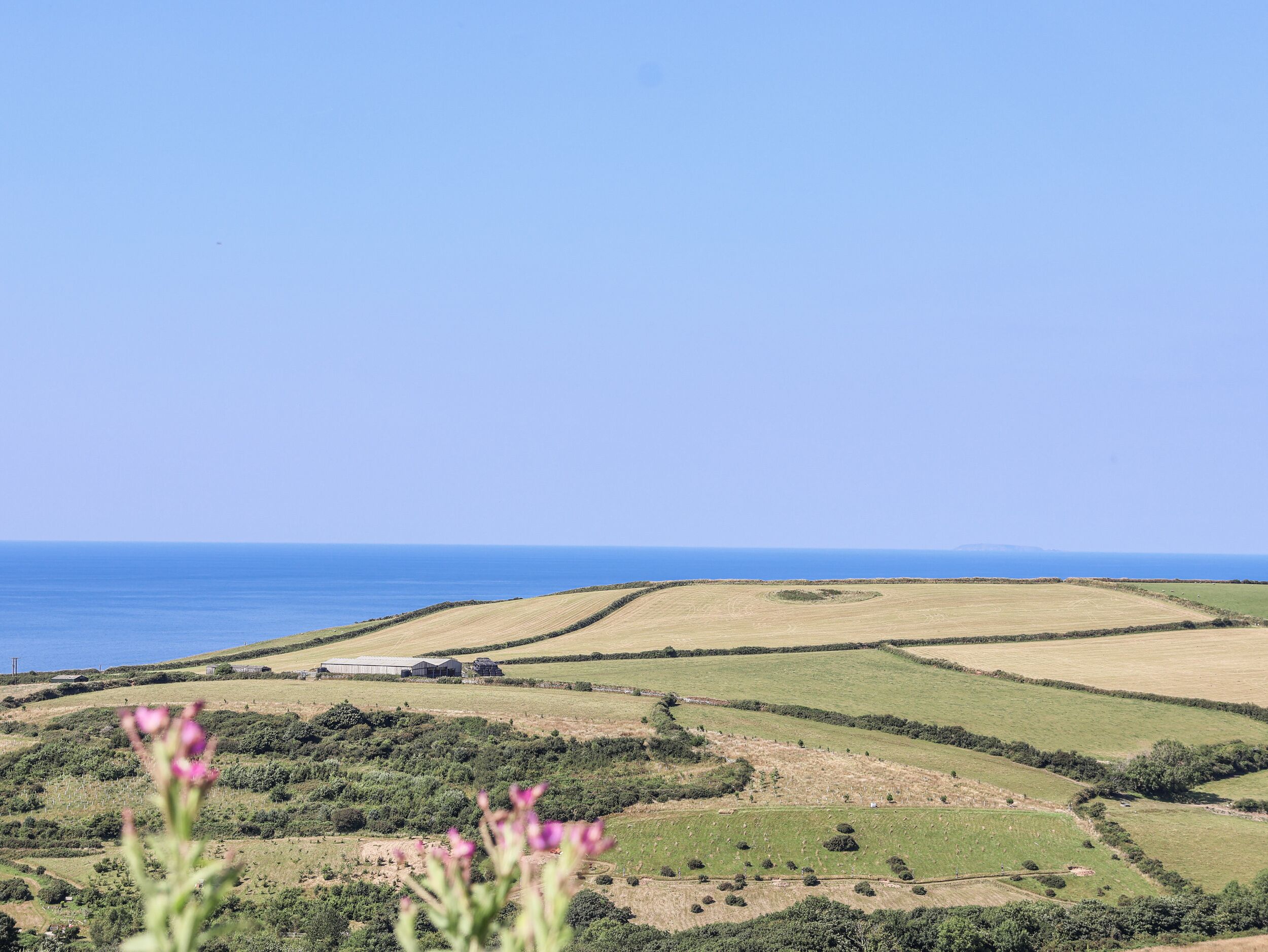 A rural landscape with rolling fields, a clear sky, and a distant view of the sea.