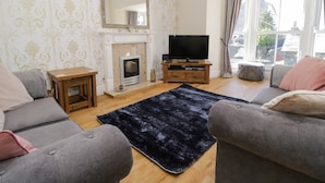 A living room with two grey sofas, a black rug, a fireplace, a television, and a large window with curtains. - Chapel Rest (Windermere)