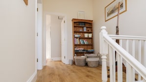 A hallway with a wooden floor, a white staircase, and a wooden bookshelf with baskets. - Chapel Rest (Windermere)