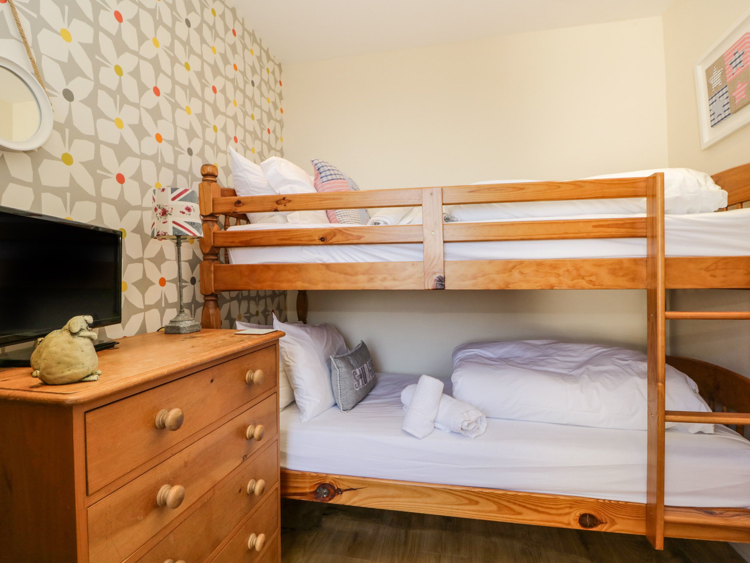 A bunk bed room with a wooden dresser, a television, and patterned wallpaper.