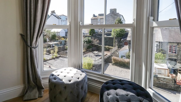 A room with a large bay window, a grey tufted ottoman, and a view of a residential area. - Chapel Rest (Windermere)
